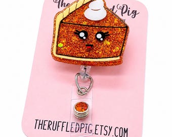 Könnte beinhalten: Ein glitzernder, orangefarbener Kürbiskuchen-Ausweishalter mit einem Kawaii-Gesicht, großen schwarzen Augen und einem kleinen Lächeln, gekrönt von einem weißen Klecks Schlagsahne. Das Acrylteil ist an einem transparenten Kunststoffband befestigt.
