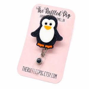 May include: A badge reel featuring a glittery black penguin with a white belly, orange feet, and a yellow beak. The penguin is attached to a retractable cord on a light pink display card printed with The Ruffled Pig and THERUFFLEDPIG.ETSY.COM.