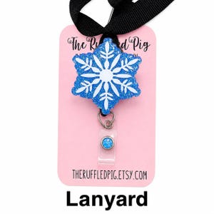 May include: A blue glittery snowflake badge reel attached to a black lanyard. The snowflake features a white center design. The badge is mounted on a light pink display card with the text The Ruffled Pig and theruffledpig.etsy.com printed on it.