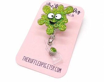 May include: A lime green glittery badge reel featuring a cartoonish blob character with large white eyes and a black smiling mouth. The badge reel is attached to a clear plastic strap with a small green glittery button, mounted on a pink display card.