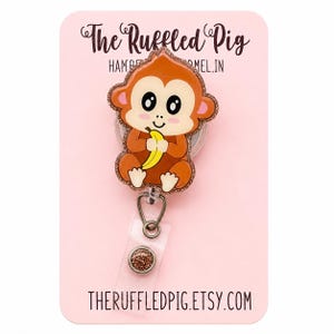 May include: A badge reel featuring a cartoon monkey holding a yellow banana. The monkey has brown fur, a light tan face, and large black eyes. The design is outlined in a copper-colored glitter border and is attached to a clear plastic strap.