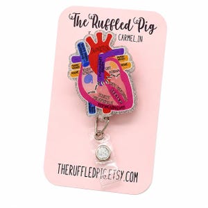 May include: A glittery acrylic badge reel featuring an anatomical heart illustration with labeled parts including the aorta, right atrium, and septum. The heart is pink, red, and purple, mounted on a light pink display card with The Ruffled Pig text.