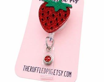 May include: A red glitter strawberry badge reel with a green glitter stem, attached to a clear plastic retractable reel. The badge reel is on a pink card with the text "The Ruffled Pig" and "THERUFFLEDPIG.ETSY.COM".