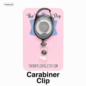 May include: A retractable badge reel with a black carabiner clip and a clear plastic strap featuring a metal snap button. The reel is mounted on a light blue glittery backing attached to a pink display card with the text The Ruffled Pig.