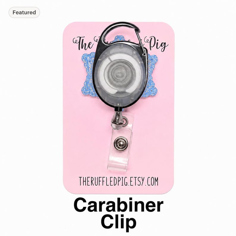 May include: A retractable badge reel with a black carabiner clip and a clear plastic strap featuring a metal snap button. The reel is mounted on a light blue glittery backing attached to a pink display card with the text The Ruffled Pig.