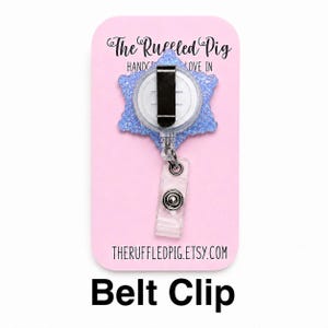 May include: A light blue glittery snowflake-shaped badge reel with a black metal belt clip and a clear plastic strap with a snap button. The item is mounted on a pink display card featuring the text The Ruffled Pig and theruffledpig.etsy.com.