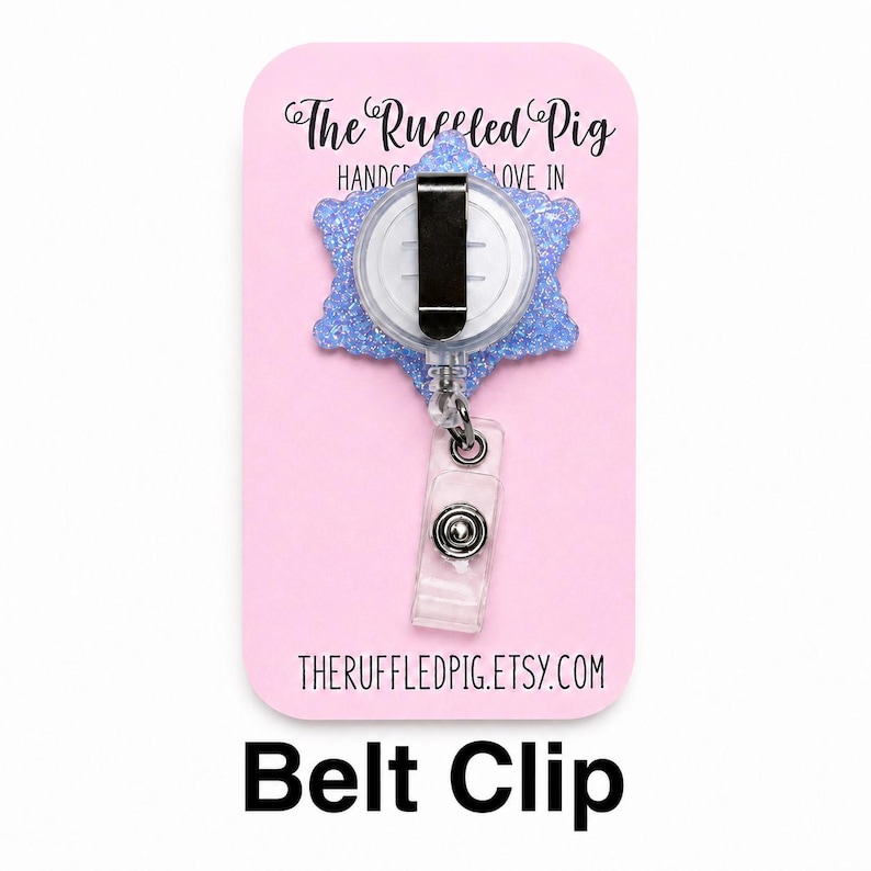 May include: A light blue glittery snowflake-shaped badge reel with a black metal belt clip and a clear plastic strap with a snap button. The item is mounted on a pink display card featuring the text The Ruffled Pig and theruffledpig.etsy.com.