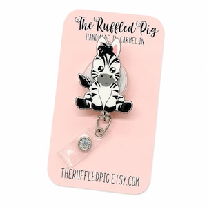 May include: A badge reel featuring a cartoon zebra with black and white stripes, large eyes, and a grey muzzle. The zebra is attached to a clear plastic strap with a silver snap button. The display card reads The Ruffled Pig, Handmade in Carmel, IN.