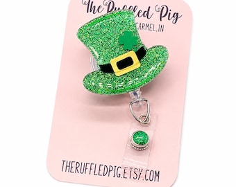 May include: A green glittery leprechaun hat badge reel with a black band, yellow buckle, and a small green shamrock accent. The badge reel is attached to a light pink display card featuring the text The Ruffled Pig and THERUFFLEDPIG.ETSY.COM.