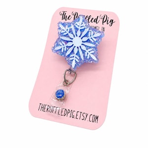 May include: A snowflake-shaped badge reel with a blue and white glitter design. The badge reel is attached to a pink card with the text "The Ruffled Pig" and "THERUFFLEDPIG.ETSY.COM".
