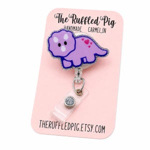 May include: A purple triceratops badge reel with a glittery border and small pink hearts on its back. The badge reel is attached to a clear plastic strap with a silver glitter button, mounted on a light pink display card labeled The Ruffled Pig.