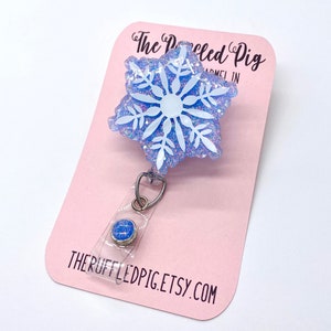 May include: A badge reel featuring a light blue snowflake shape with white snowflake detailing and iridescent glitter embedded in the resin. The badge reel is attached to a light pink display card with the text The Ruffled Pig and THERUFFLEDPIG.ETSY.COM.