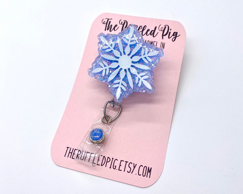 May include: A badge reel featuring a light blue snowflake shape with white snowflake detailing and iridescent glitter embedded in the resin. The badge reel is attached to a light pink display card with the text The Ruffled Pig and THERUFFLEDPIG.ETSY.COM.