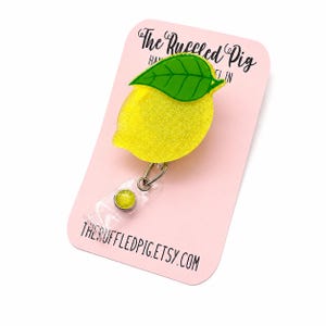 May include: A yellow glittery lemon-shaped badge reel with a green leaf accent, attached to a clear plastic strap with a small yellow circular button. The item is mounted on a light pink display card featuring the text The Ruffled Pig.