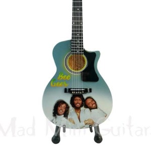 Puede incluir: Una guitarra acústica azul con una golpeador negro y una imagen de los Bee Gees en el cuerpo. La guitarra está en un soporte.