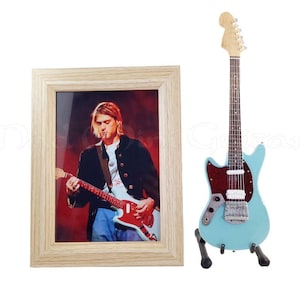 Puede incluir: Una fotografía enmarcada de un músico tocando una guitarra roja, junto a una guitarra eléctrica en miniatura de color azul claro con un golpeador marrón. La fotografía está en un marco de madera clara. La guitarra en miniatura está sobre un soporte negro.