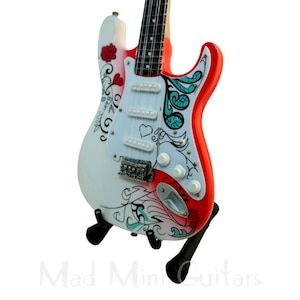 Könnte beinhalten: Eine Miniatur-E-Gitarre mit weißem Korpus, roter Umrandung und schwarzem Hals. Die Gitarre ist mit floralen und Herzmotiven in Rot und Türkis verziert. Sie wird auf einem schwarzen Ständer präsentiert. Der Text "Mad Mini Guitars" ist unten sichtbar.