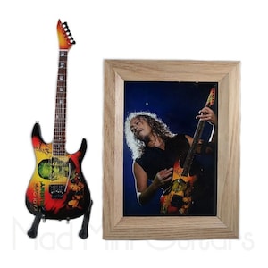 Pu&ograve; includere: Una replica in miniatura di una chitarra elettrica nera e rossa con un design grafico colorato. La chitarra &egrave; su un supporto e si trova davanti a una foto incorniciata di un musicista che suona una chitarra simile.