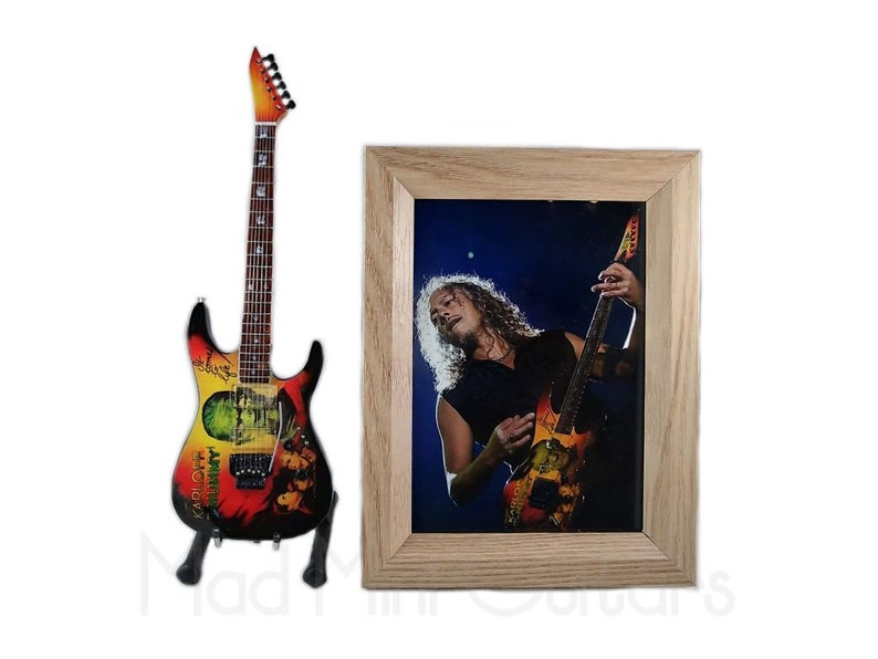 Pu&ograve; includere: Una replica in miniatura di una chitarra elettrica nera e rossa con un design grafico colorato. La chitarra &egrave; su un supporto e si trova davanti a una foto incorniciata di un musicista che suona una chitarra simile.