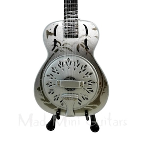 May include: A silver-colored miniature resonator guitar with a black stand. The guitar has a round sound hole with a decorative design and a palm tree pattern on the body.