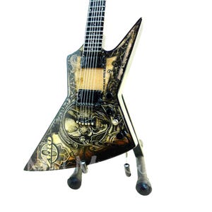 May include: A detailed, miniature electric guitar with a unique design. The guitar features a cream-colored body with intricate black and brown artwork, including skulls and ornate patterns. It is displayed on a small stand against a white background.