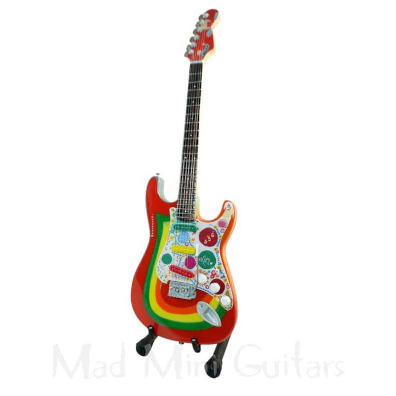 May include: A colorful electric guitar with a red body, rainbow stripes, and various decorative elements. The guitar is supported by a black stand. The headstock is red, and the fretboard is dark. The guitar has the text "Mad Mind Guitars" at the bottom.