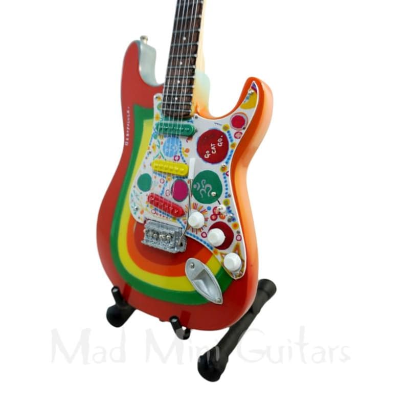 May include: A miniature electric guitar with a vibrant rainbow design on a black stand. The guitar features a red body with yellow, orange, and green accents. The pickguard is white with colorful circles and shapes. The text "Mad Mini Guitars" is visible.