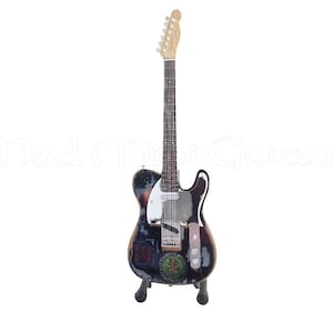 Może przedstawiać: Czarno-biała gitara elektryczna z wytartym wykończeniem i różnymi naklejkami na korpusie. Gitara stoi na stojaku i ma biały pickguard.