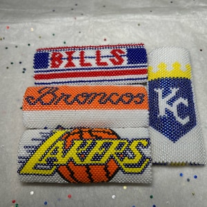 May include: Four beaded keychains with different sports team logos. The keychains are white with blue, red, yellow, and orange beads. The logos are for the Bills, Broncos, Lakers, and Kansas City Chiefs.