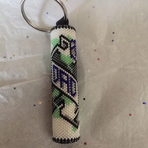 May include: A white, green, blue, and black beaded keychain with a silver key ring. The beads are arranged in a pattern that resembles a ribbon.