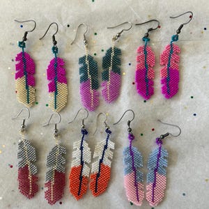 May include: A collection of beaded feather earrings in various colors, including pink, blue, white, and orange. Each earring features a unique design with silver-tone hooks. The earrings are arranged on a light-colored surface.