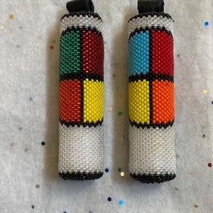 May include: Two beaded cylindrical accessories with a black leather loop at the top. Each cylinder is divided into four colored sections: green, red, yellow, and orange, and blue, red, yellow, and orange. The base is white with black and silver accents.