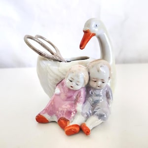 May include: A white ceramic swan figurine with an orange beak and a small opening for holding items. Two small figurines of children in pink and purple clothing are seated at the base. Silver scissors are placed in the opening.