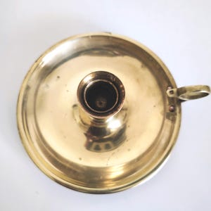 Puede incluir: Un candelero de latón vintage. Presenta una base circular, un receptáculo central para la vela y un pequeño asa. El latón tiene un acabado pulido y reflectante. El diseño es sencillo y elegante, adecuado para una sola vela.