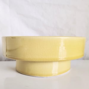 May include: A pale yellow ceramic bowl with a glossy finish and a visible crackle glaze pattern. The bowl features a wide, circular upper section that tapers down to a smaller, solid pedestal base, creating a tiered, elevated silhouette.
