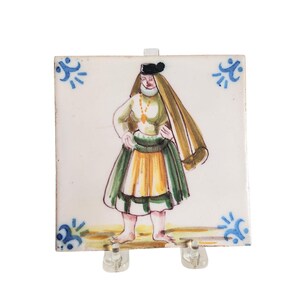 Puede incluir: Azulejo cerámico cuadrado con una ilustración pintada a mano de una persona con vestimenta tradicional. La figura lleva una falda verde y amarilla, una blusa verde claro y un sombrero negro. Acentos florales azules adornan las esquinas.