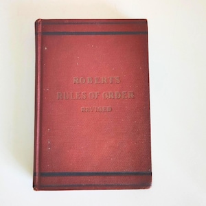 May include: A vintage red book titled "Roberts Rules of Order Revised." The book has gold lettering and dark blue stripes at the top and bottom. The book is on a white background.