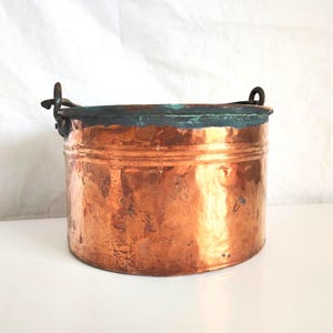 Puede incluir: Un cubo de cobre con forma cilíndrica y dos asas pequeñas. El cubo tiene un color marrón rojizo intenso con una pátina ligeramente envejecida. El borde del cubo tiene un tono verde oscuro, lo que añade contraste al cobre.