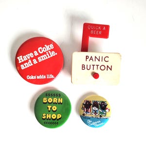 May include: A collection of vintage buttons and a small sign. The buttons include a red one with "Have a Coke and a smile" and a green one that says "BORN TO SHOP". The sign reads "PANIC BUTTON" and "QUICK A BEER".