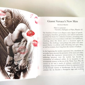 May include: A book page featuring a black and white photograph of a muscular person with red lipstick kiss marks overlaid on the image. The text on the opposite page is titled Gianni Versace's New Men by Richard Martin and discusses fashion.