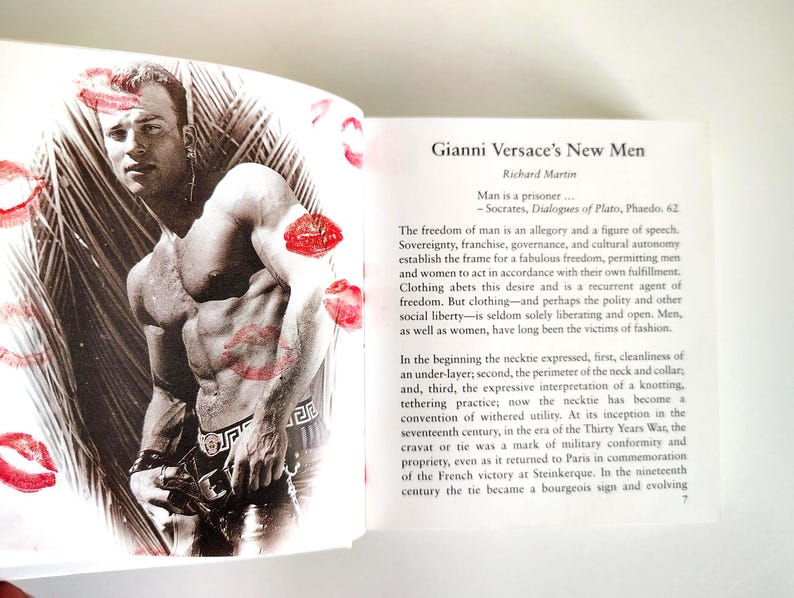 May include: A book page featuring a black and white photograph of a muscular person with red lipstick kiss marks overlaid on the image. The text on the opposite page is titled Gianni Versace's New Men by Richard Martin and discusses fashion.