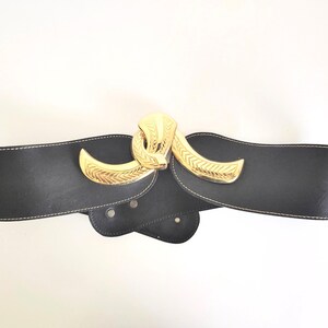 May include: A wide black belt featuring a prominent gold-tone metal buckle with a textured, curved design. The belt is constructed from a smooth black material with visible white stitching along the edges and includes a series of adjustment holes.
