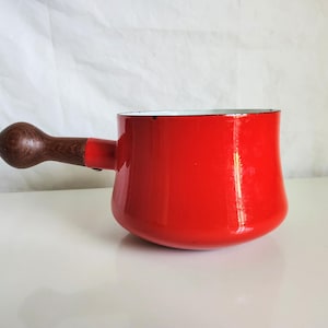 May include: A bright red enameled metal saucepan with a smooth, glossy finish and a rounded, dark brown wooden handle. The pot features a wide, cylindrical body with a slightly curved base and a clean, simple design for stovetop cooking.