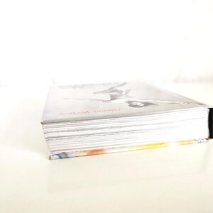 May include: A hardcover book with a silver-toned cover featuring a black and white illustration of a figure and the red text Gianni Versace. The book is positioned at an angle, showing the white pages of the text block and a black spine.