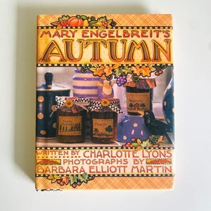 May include: A book cover titled "Mary Engelbreit's Autumn" with illustrations of jars of preserves, a black bird, and fall foliage. The cover features text by Charlotte Lyons and photographs by Barbara Elliott Martin.