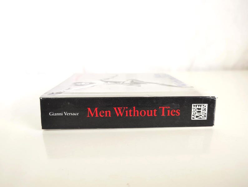 May include: A hardcover book titled Men Without Ties by Gianni Versace, featuring a black spine with red text and the Abbeville Press logo. The book is positioned horizontally, showing the side edge with clear white lettering and branding.