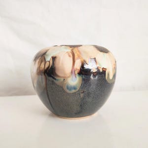 May include: A ceramic vase with a rounded shape. The vase has a dark gray base with a glossy finish. The top portion features a blend of beige, brown, and light blue hues, creating a unique, dripped effect. The vase sits on a white surface.