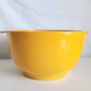 May include: A bright yellow mixing bowl with a smooth, solid-colored finish. The bowl features a rounded body, a slightly raised circular base, and a small, integrated handle tab on one side for easier gripping and handling during kitchen tasks.