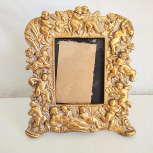 May include: Ornate gold-colored picture frame with cherub and floral relief details. The frame has a rectangular opening with a dark backing and a piece of tan paper. The frame is standing upright on a white surface.