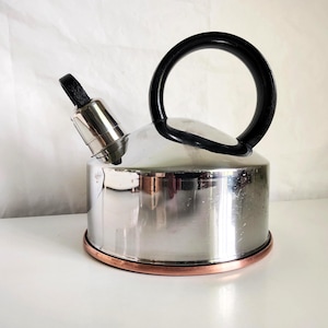 May include: A polished stainless steel stovetop kettle with a rounded body, a black arched handle, and a silver spout with a black tip. The base of the kettle features a distinct copper-colored trim ring around the bottom edge of the metal body.